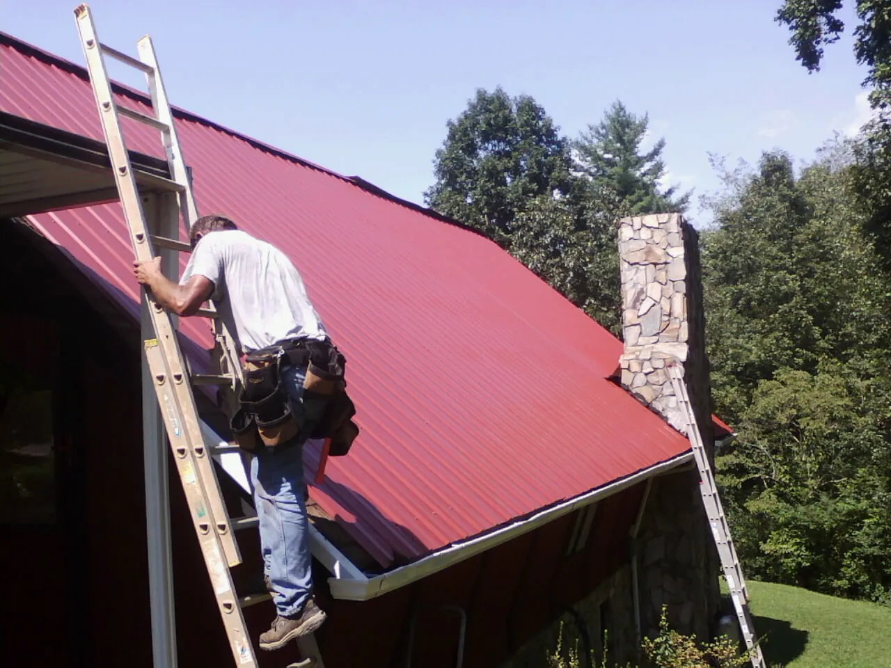 Roofer on a ladder beside a red metal roof and stone chimney — Soffit & Fascia Repair by Roofing Brothers in Andalusia